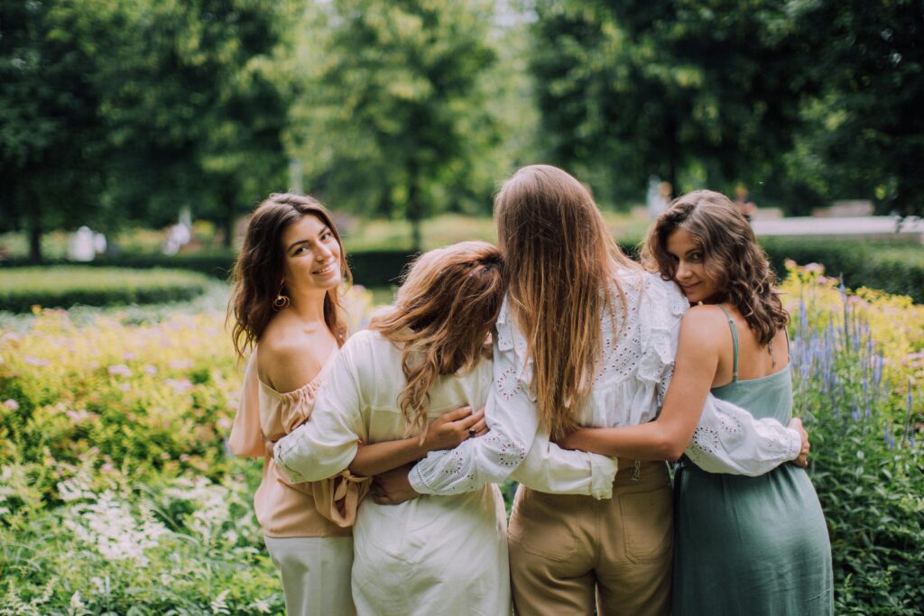 Four women enjoying a sunny day together in a green park, embracing each other and smiling.