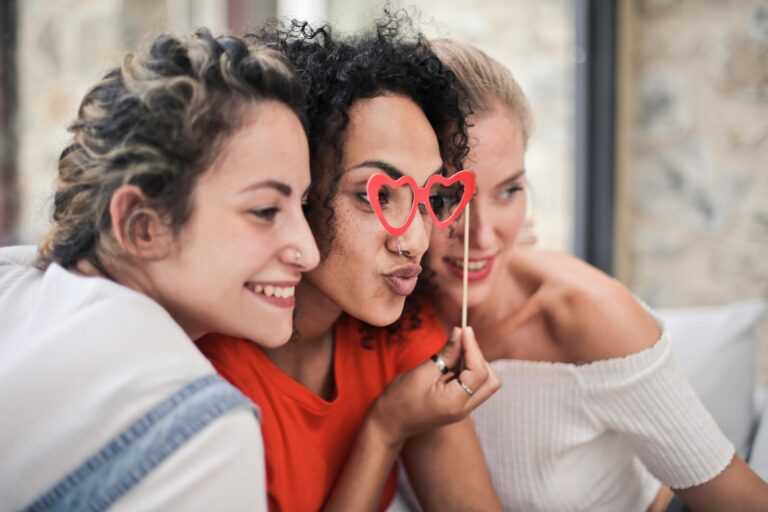 Three women enjoying a playful moment with heart-shaped glasses, symbolizing friendship and fun. Making new friends.